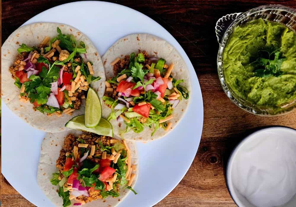 Overhead view of three Tofu Street Tacos on a white plate with lime wedges, served with bowls of guacamole and sour cream.
