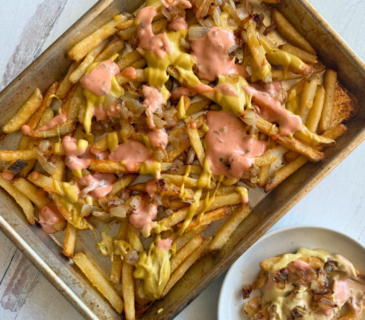 A sheet pan of vegan animal style fries topped with cashew cheese sauce, special sauce, and browned onions, with a served plate on the side.