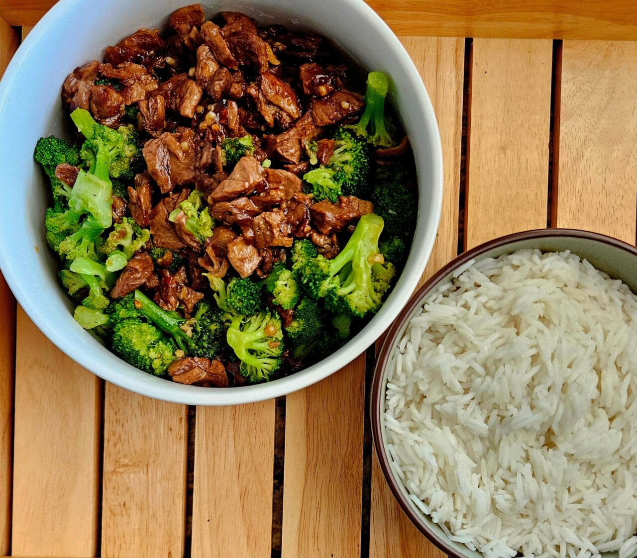 Vegan Beef and Broccoli with plant-based steak bites and broccoli in a savory sauce in a white bowl, next to a bowl of white rice on a wooden slatted tray.