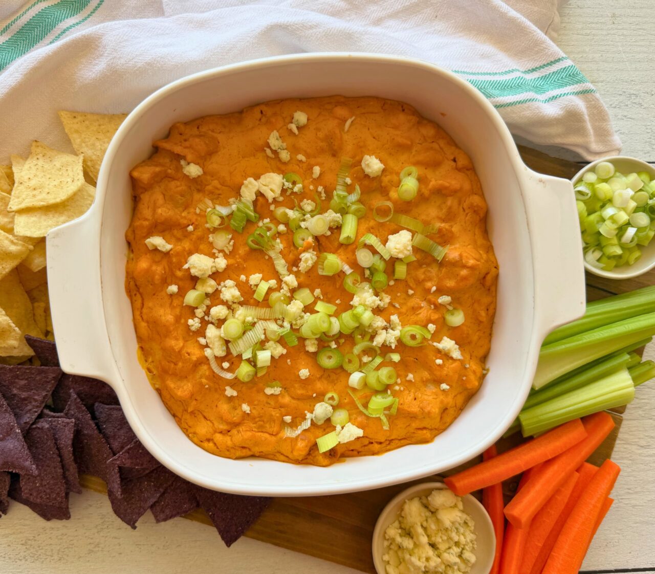 Vegan buffalo chicken dip topped with green onions and vegan blue cheese crumbles in a white baking dish, surrounded by tortilla chips, celery, and carrot sticks.