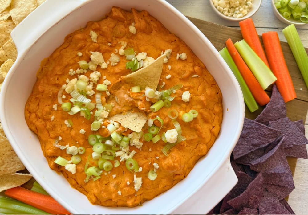 Close-up of creamy vegan buffalo chicken dip with tortilla chips dipped in, topped with green onions and vegan blue cheese crumbles.