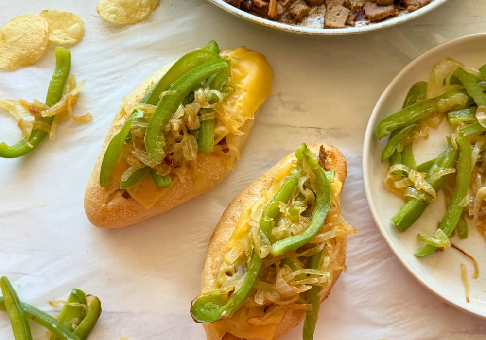 Two vegan Philly cheesesteaks topped with melted vegan provolone, caramelized onions, and green peppers, with a skillet of vegan beef and mushrooms and a plate of extra onions and peppers nearby.